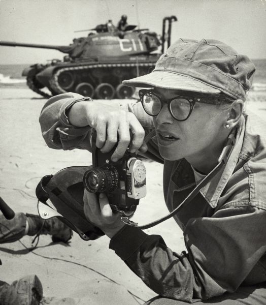 Dickey Chapelle, photographer, on the same Milwaukee beach where she learned to swim as a young girl. She was covering "Operation Inland Seas" celebrating the opening of the St. Lawrence Seaway. She is holding her camera and there is a tank in the background. This is her favorite photograph of herself at work.