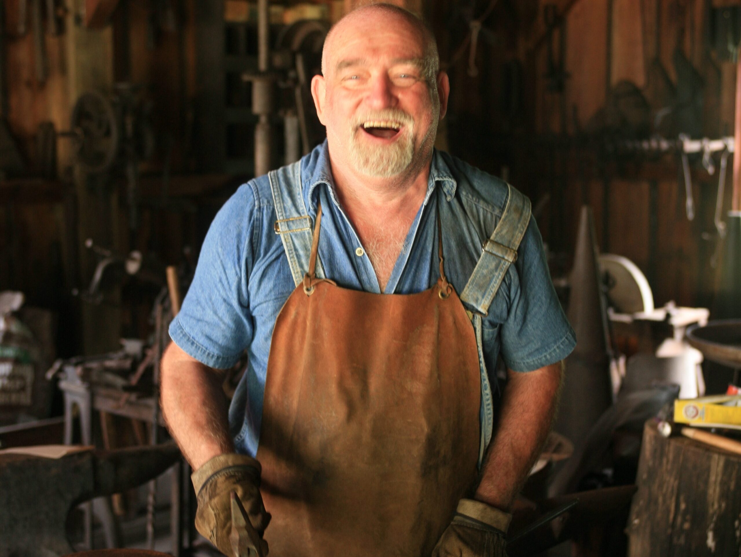 A blacksmith, man, laughs, happily at the camera while working a piece of metal during his employment. 