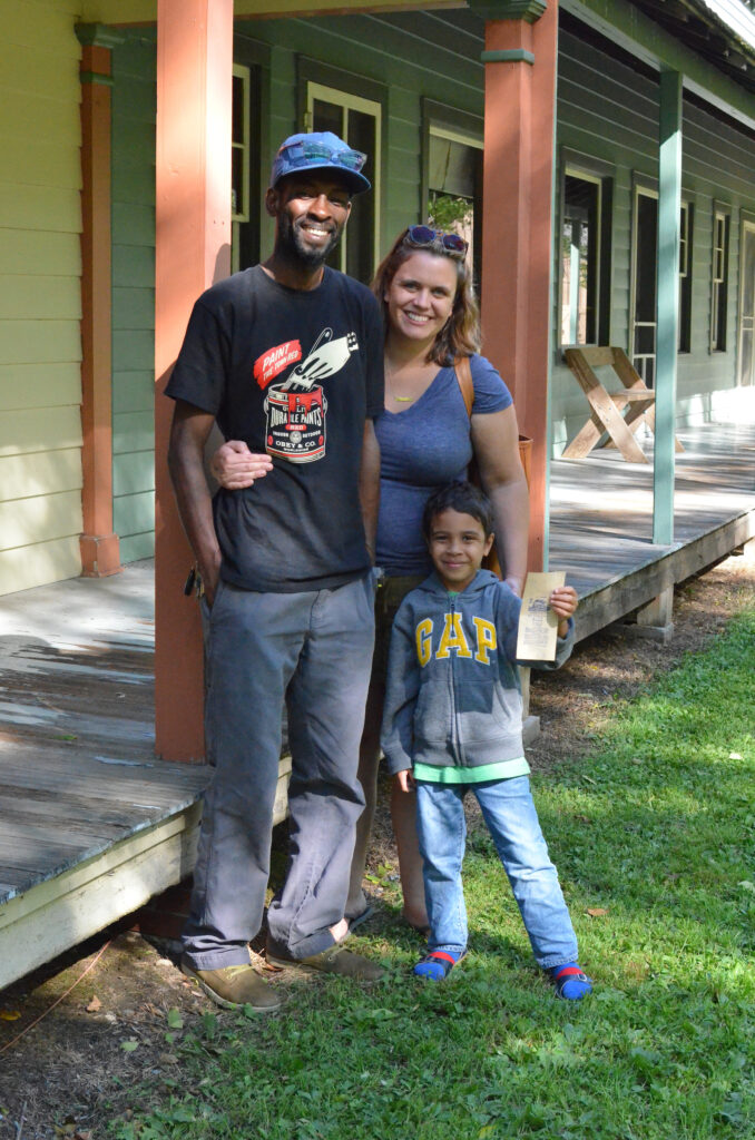 A couple, a black man wearing a black shirt and a white woman wearing a blue shirt, posing for a photo with their son.