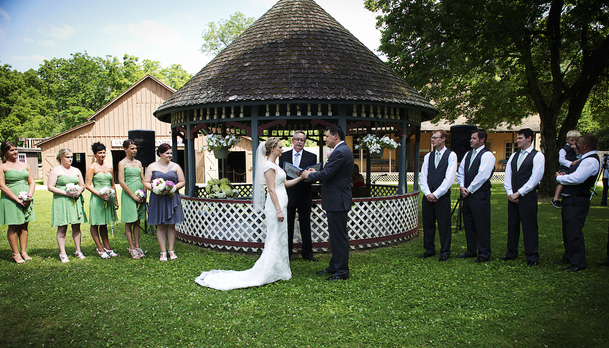 A couple actively getting married by a gazebo with the bridesmaids and groomsmen nearby