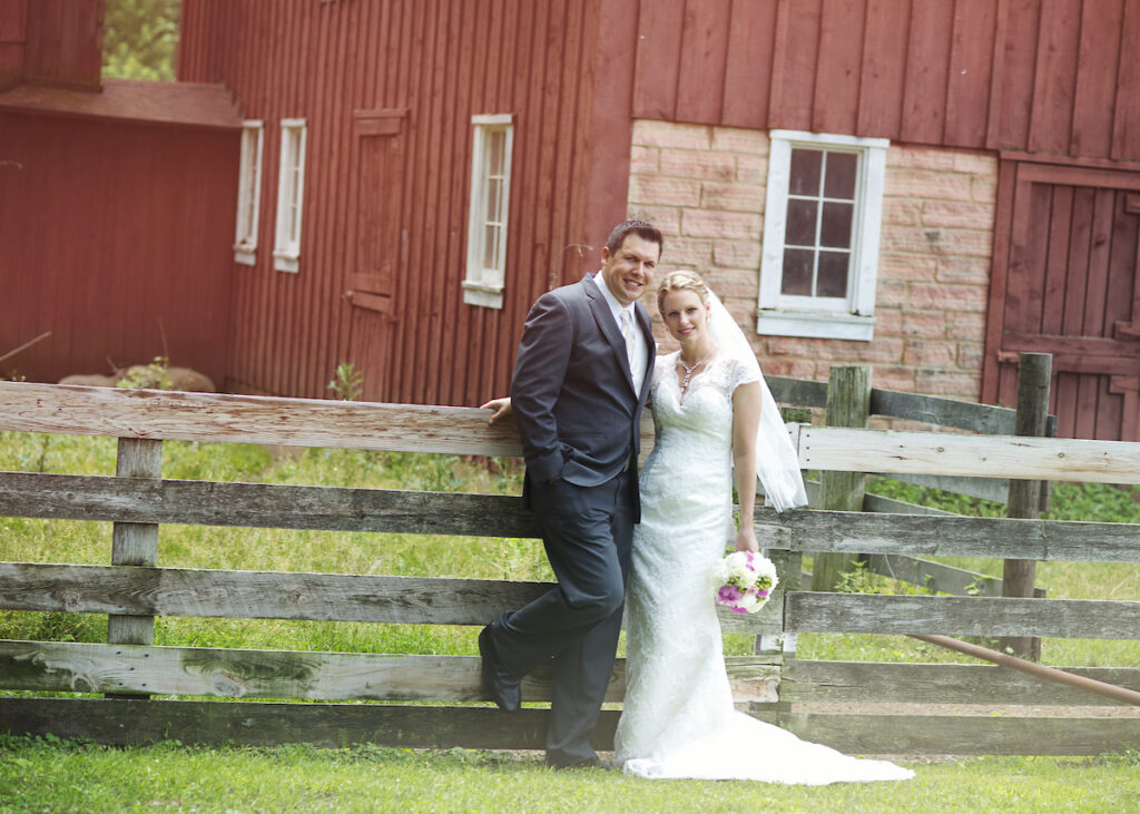A newly wed couple posing and smiling in front of a barn and the fence