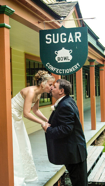 A newly wed couple, wearing a wedding dress and suit, kissing under a "Sugar Bowl Confectionery" sign.